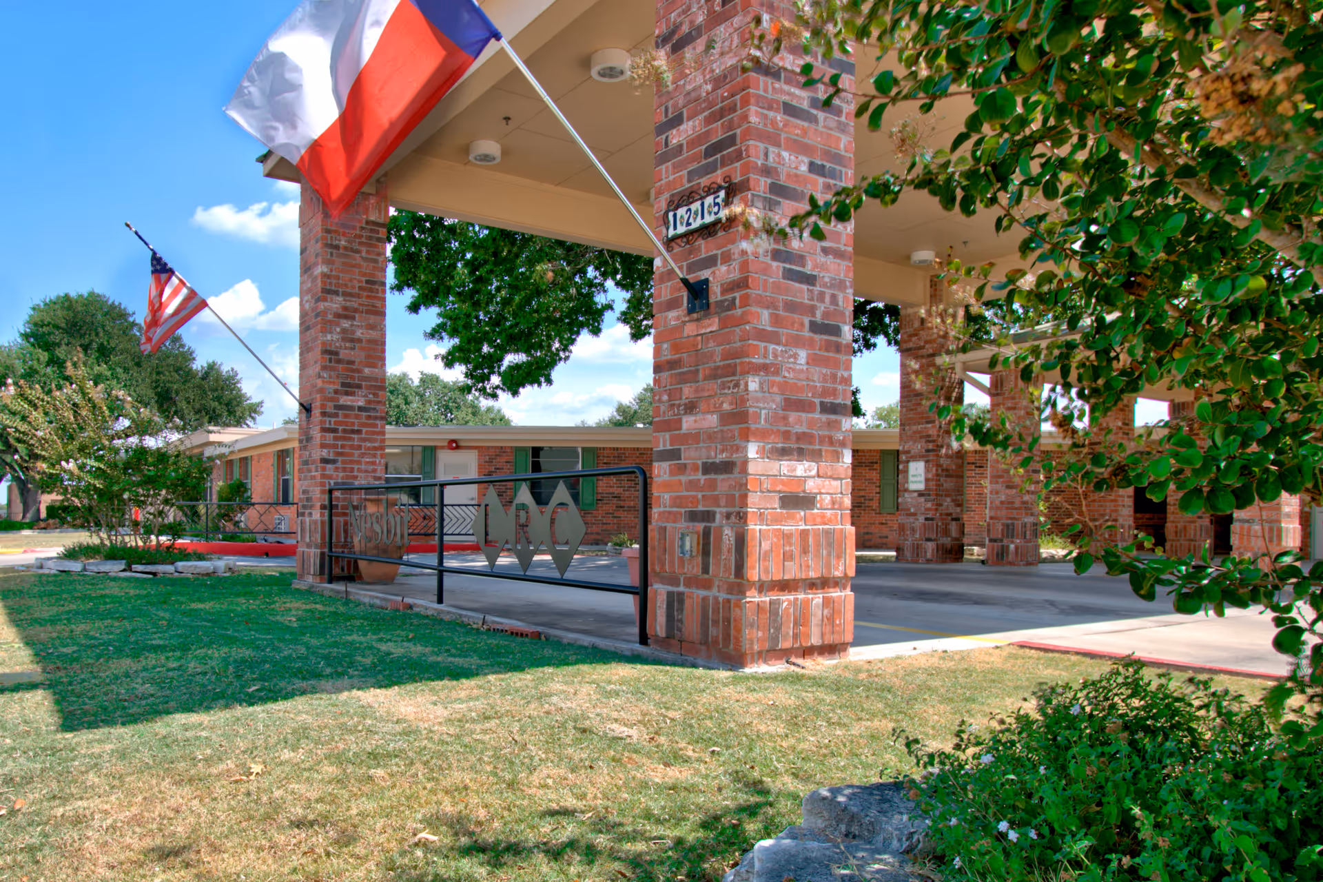 Exterior view of Nesbit Living & Recovery Center showing a brick entrance structure with two flags, an American flag and a Texas state flag, mounted on poles. The building is single-story with a brick facade, surrounded by green grass and trees under a blue sky with some clouds.