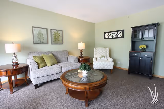 A cozy living room with a light gray sofa adorned with two green pillows, a white patterned armchair, a round wooden coffee table with a glass top and a small plant, two wooden side tables each with a lamp, two framed botanical prints on the wall, and a black cabinet with glass doors and a small yellow flower arrangement inside. The walls are painted light green and the floor is carpeted.