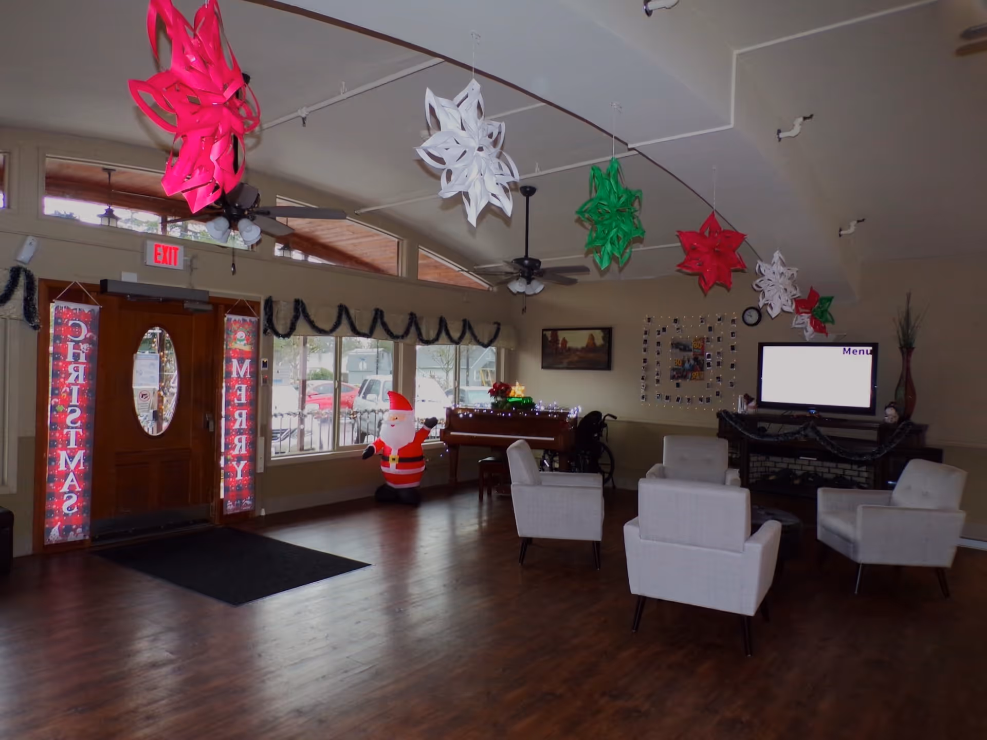 A common area decorated for Christmas with hanging red, white, and green paper snowflakes from the ceiling. There is a wooden door with glass oval window and Christmas banners on either side. An inflatable Santa Claus figure stands near the windows. Four white armchairs are arranged around a small table on a wooden floor. A piano decorated with Christmas lights and a TV screen displaying a menu are visible in the background.