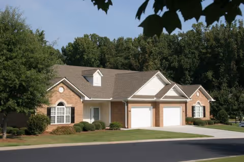 A single-story brick residential building with two white garage doors, white trim, and black shutters. The building is surrounded by well-maintained green lawns, bushes, and trees, with a clear blue sky in the background.