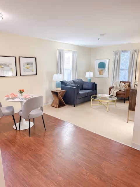 A bright and cozy living room and dining area with light-colored walls and wood flooring. The living room features a dark gray sofa, a brown armchair with a throw blanket, two side tables with lamps, a round glass coffee table with a gold frame, and two windows with light curtains. The dining area has a small round white table set with pink napkins and flowers, surrounded by four light gray upholstered chairs. Two framed abstract artworks hang on the wall above the dining table.