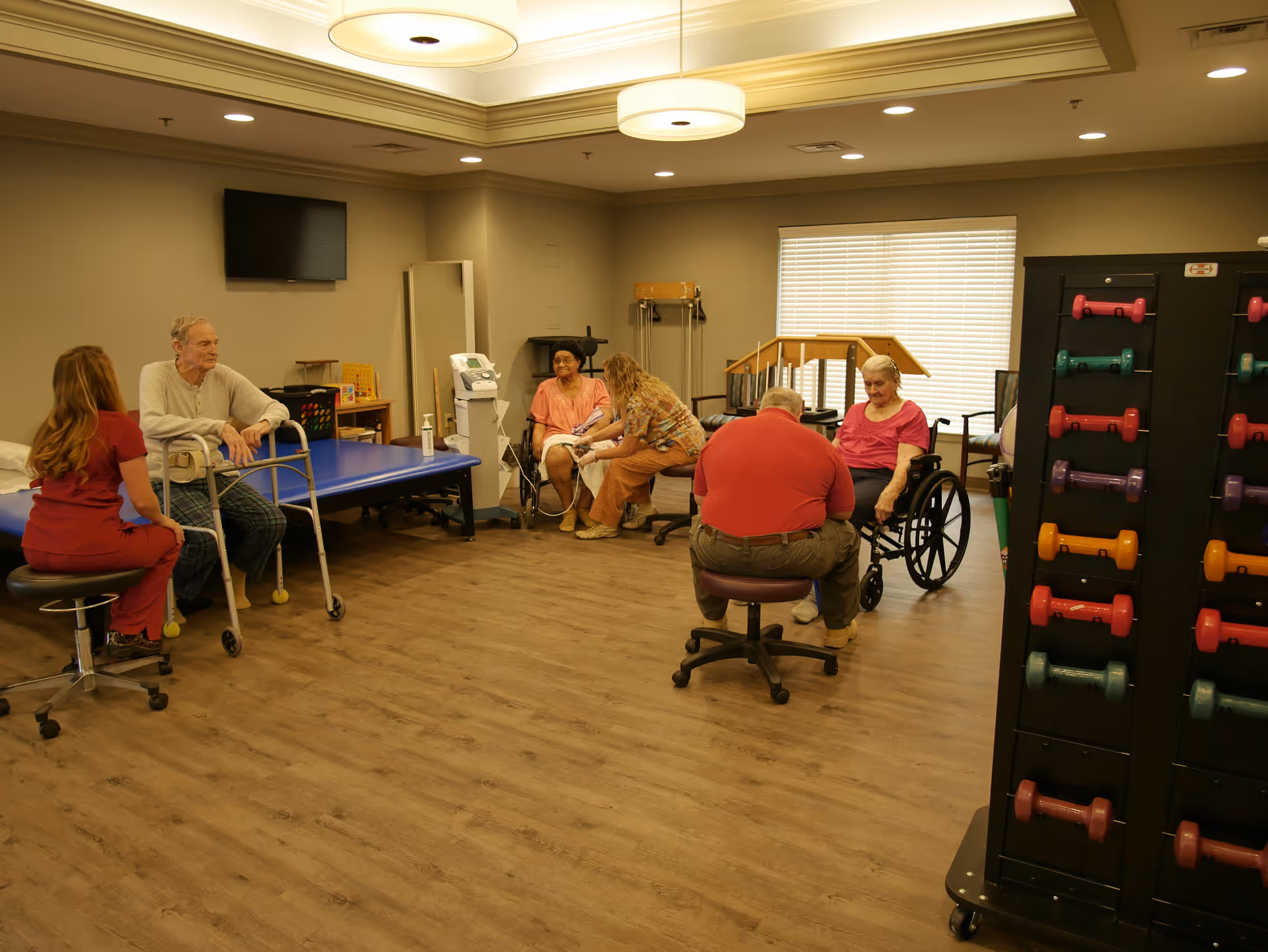 A rehabilitation room with elderly patients and caregivers. Two elderly individuals are seated, one using a walker and another in a wheelchair. Caregivers are attending to them. The room has exercise equipment including a rack of colorful dumbbells, a therapy table, and a large window with blinds. The floor is wooden and the lighting is warm.