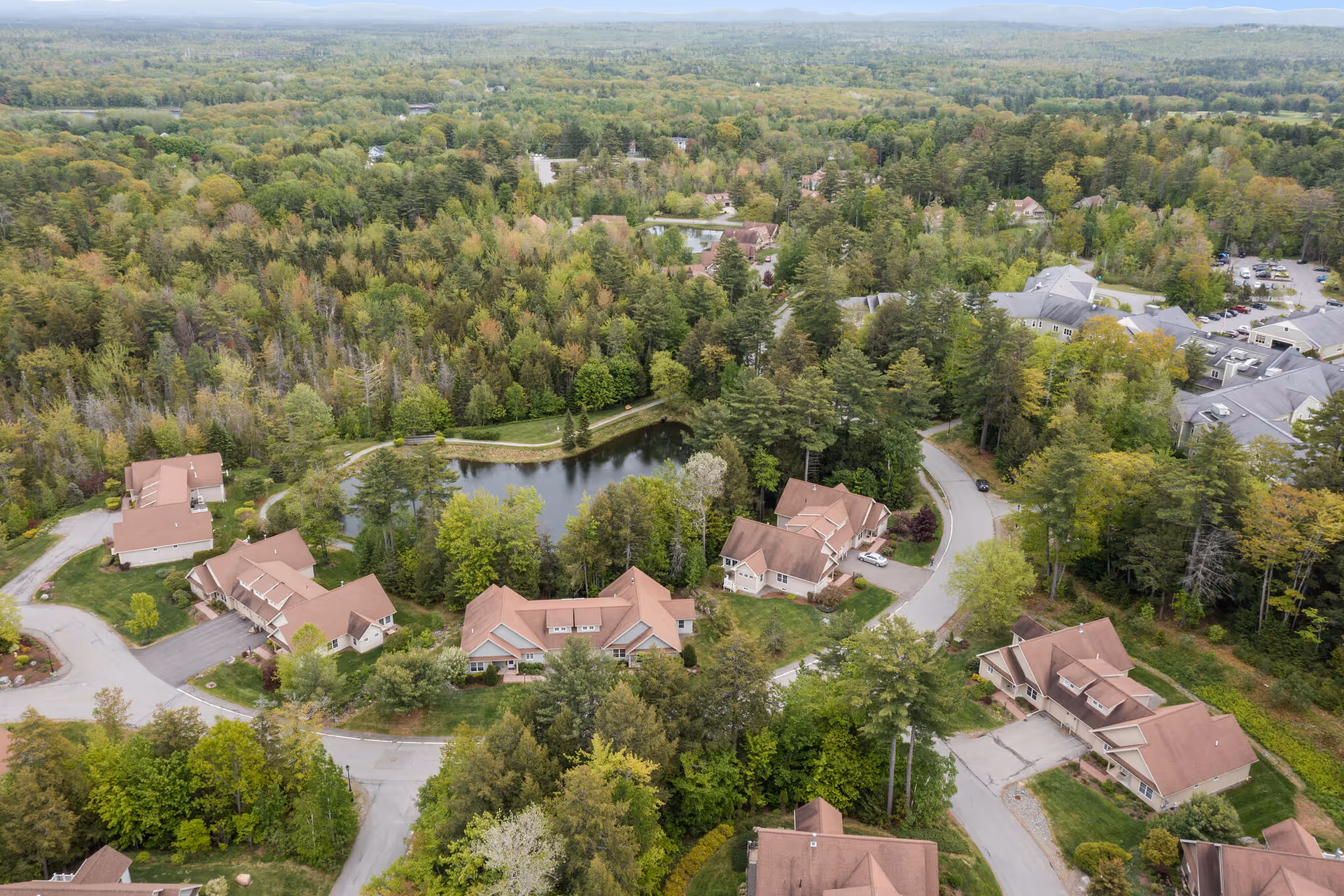 Aerial view of Dirigo Pines senior living facility surrounded by dense green forest. The image shows multiple residential buildings with brown roofs arranged around a small pond and winding roads. The landscape is lush with trees and greenery extending into the distance.