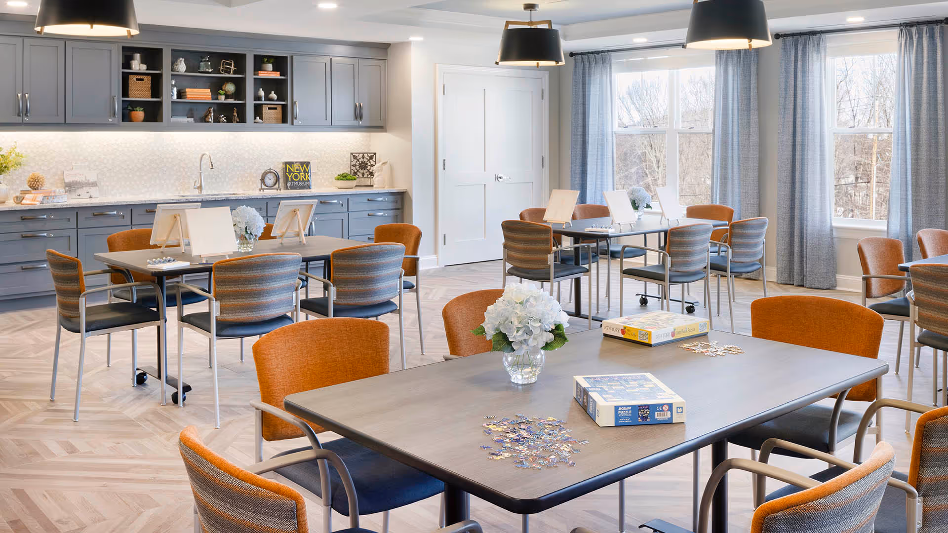 Bright communal dining and activity room with multiple tables, orange-and-gray chairs, board games, and cabinetry along the back wall.