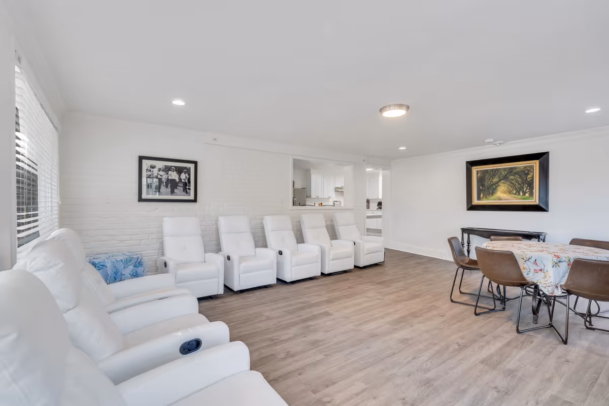 A bright and spacious living room with white walls and wood flooring. The room features a row of six white recliner chairs along one wall, a dining table with six brown chairs covered with a floral tablecloth, a framed black and white photo on the brick wall, and a framed painting of a tree-lined path on the opposite wall. A pass-through window opens to a kitchen area in the background.