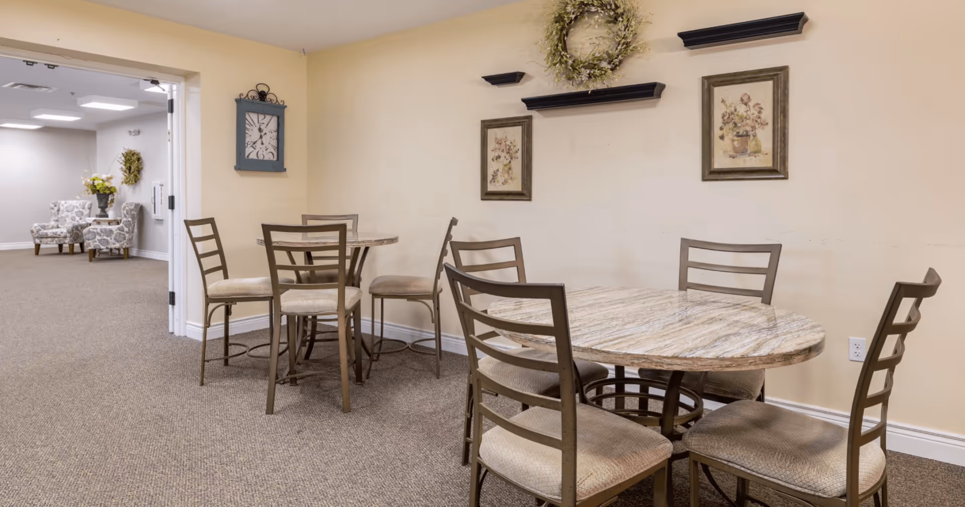 A small dining area with two marble-top tables and six chairs with beige cushions. The walls are light yellow with two framed floral pictures and a decorative wreath above the tables. In the background, there is an open doorway leading to another room with two patterned armchairs and a small table with a flower arrangement.