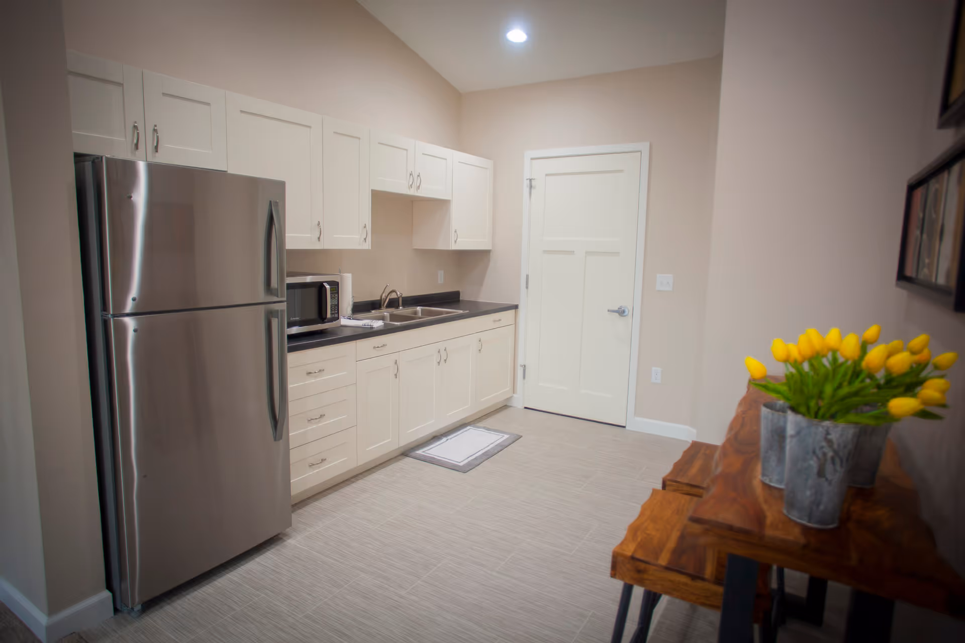A clean and modern kitchen area with white cabinets, a stainless steel refrigerator, a microwave, a sink, and a wooden table with a metal vase holding yellow tulips. The walls are painted beige and the floor has light-colored tiles.