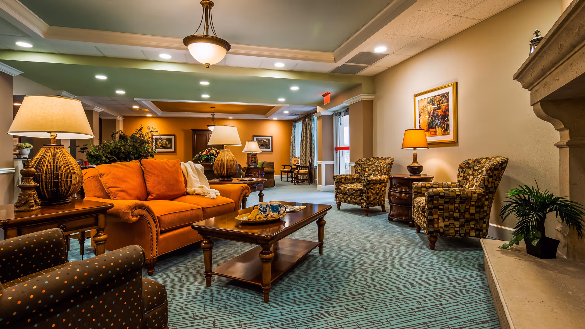 A cozy living room area in a senior living facility featuring an orange sofa, patterned armchairs, wooden coffee and side tables with lamps, decorative plants, framed artwork on the walls, and a fireplace on the right side.