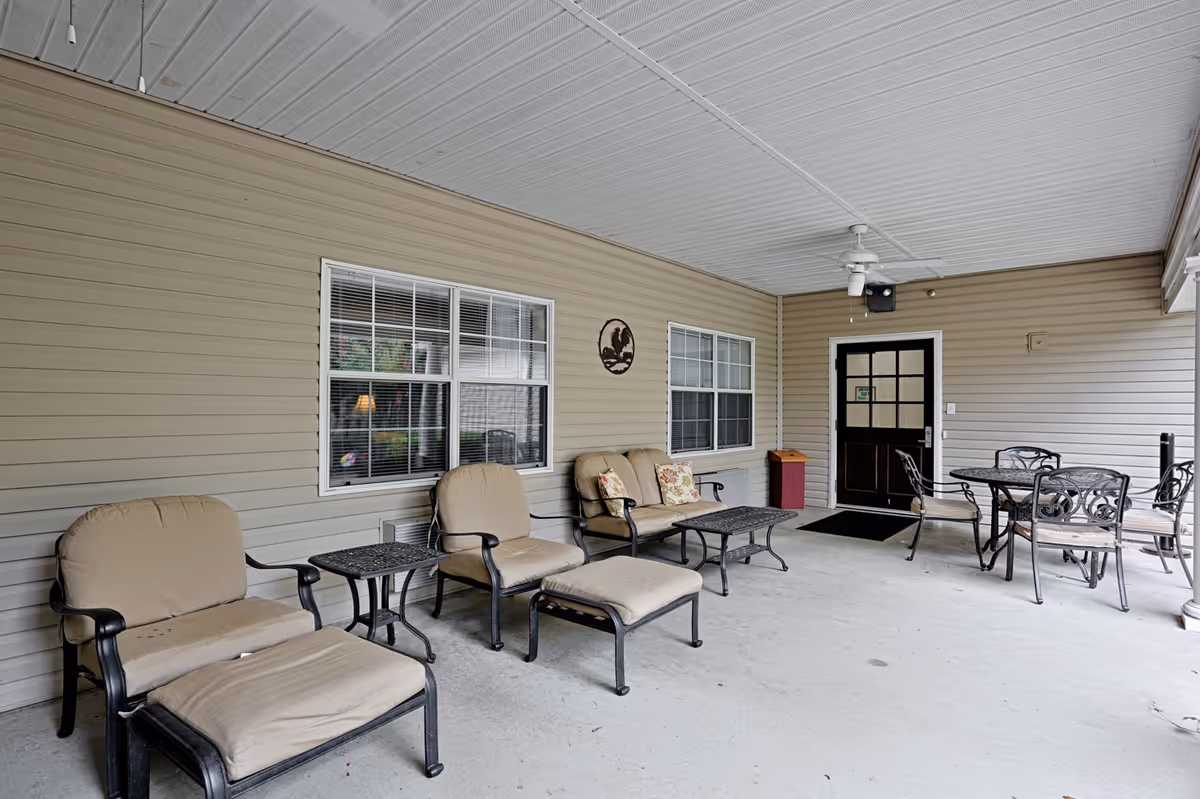 Covered outdoor patio area with beige cushioned chairs, a loveseat with floral pillows, a small side table, a coffee table, and a round dining table with four chairs. The patio has beige siding walls, two windows, a ceiling fan, and a black door with glass panels.