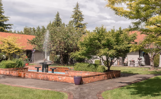 Courtyard featuring a brick fountain and raised planters surrounded by paths, trees, and single-story buildings.