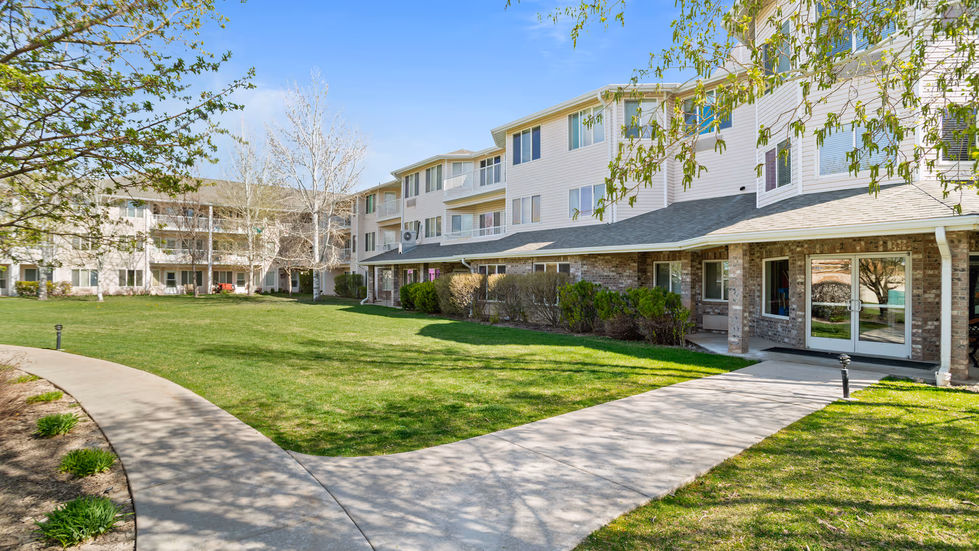 Exterior view of a multi-story senior living facility with a well-maintained green lawn, a curved concrete walkway, and trees with budding leaves under a clear blue sky.