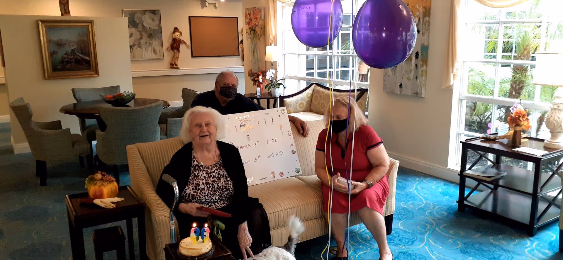An elderly woman sitting on a beige couch celebrating her 98th birthday with a small cake with candles in front of her. She is accompanied by two other people, one man and one woman, both wearing face masks. There are two large purple balloons tied to the couch. The room has blue carpet, a round table with chairs, paintings on the walls, and large windows letting in natural light.