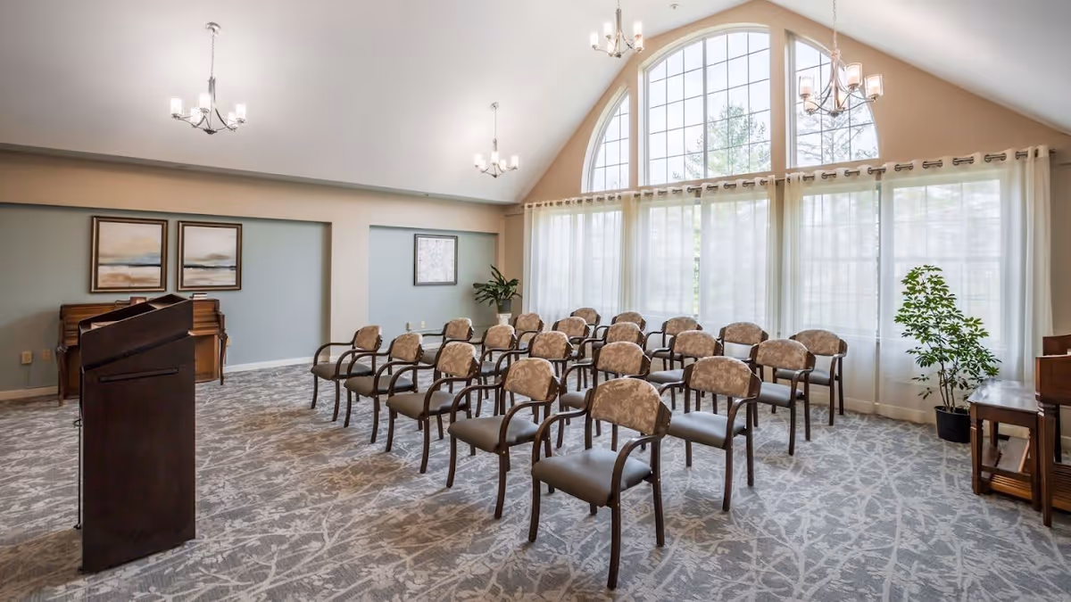 A bright and spacious meeting or presentation room with rows of upholstered chairs arranged facing a wooden podium. The room features large arched windows with sheer curtains allowing natural light to fill the space. There are chandeliers hanging from the vaulted ceiling, framed artwork on the walls, a piano, and a potted plant near the windows.