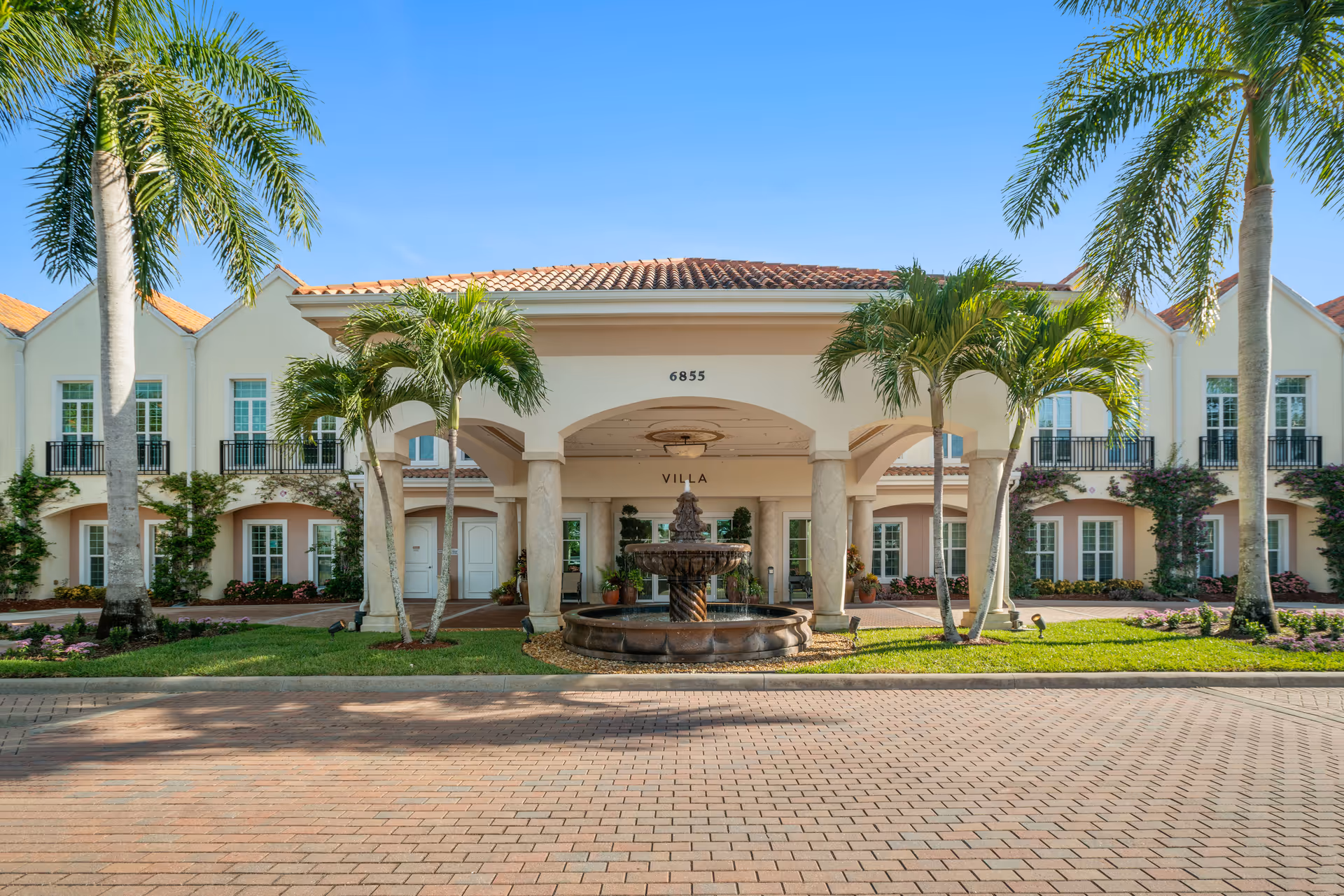 Front exterior view of a senior living facility named Villa at Terracina Grand with a tiled roof, palm trees, a central covered entrance supported by columns, and a decorative fountain in front.