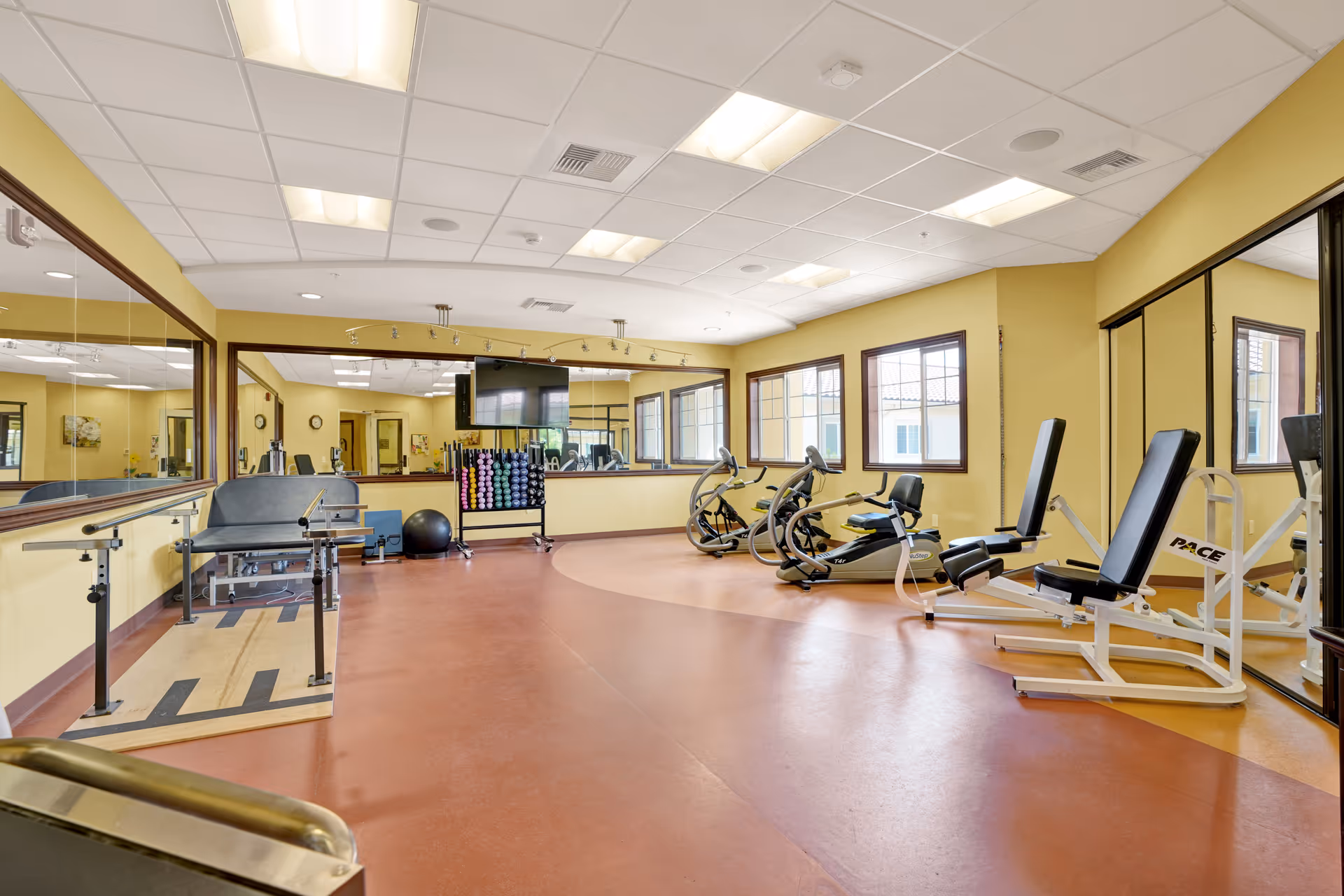A spacious fitness room with exercise equipment including stationary bikes, weight machines, and a rack of colorful dumbbells. The room has large mirrors on the walls, yellow painted walls, and several windows letting in natural light. The floor is a reddish-brown color with a lighter circular section in the middle.
