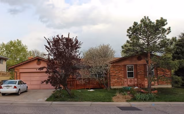 A brick, single-story house with pink garage doors, a driveway, and trees in the front yard.