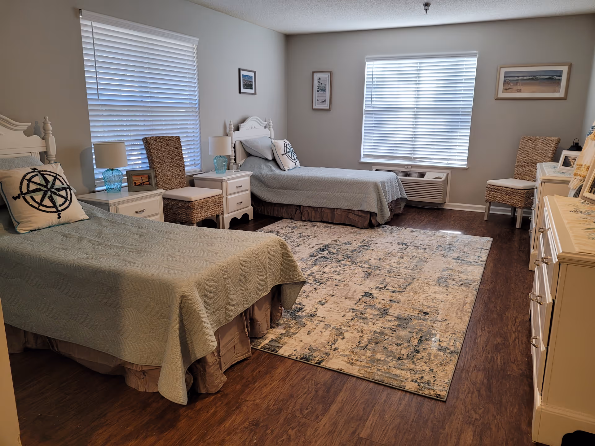 A bright bedroom with two twin beds covered in light blue quilts, each with a decorative pillow featuring a compass design. Between the beds is a white nightstand with a blue glass lamp and a framed photo. The room has two windows with white blinds, a patterned area rug on wooden flooring, two wicker chairs, and a white dresser with framed pictures on top. The walls are decorated with three framed pictures.