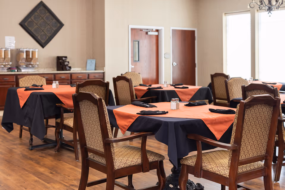 Dining room with several tables covered in black tablecloths and orange overlays, each set with black napkins and salt and pepper shakers. Wooden chairs with patterned upholstery surround the tables. In the background, there is a counter with cereal dispensers and a decorative wall piece.