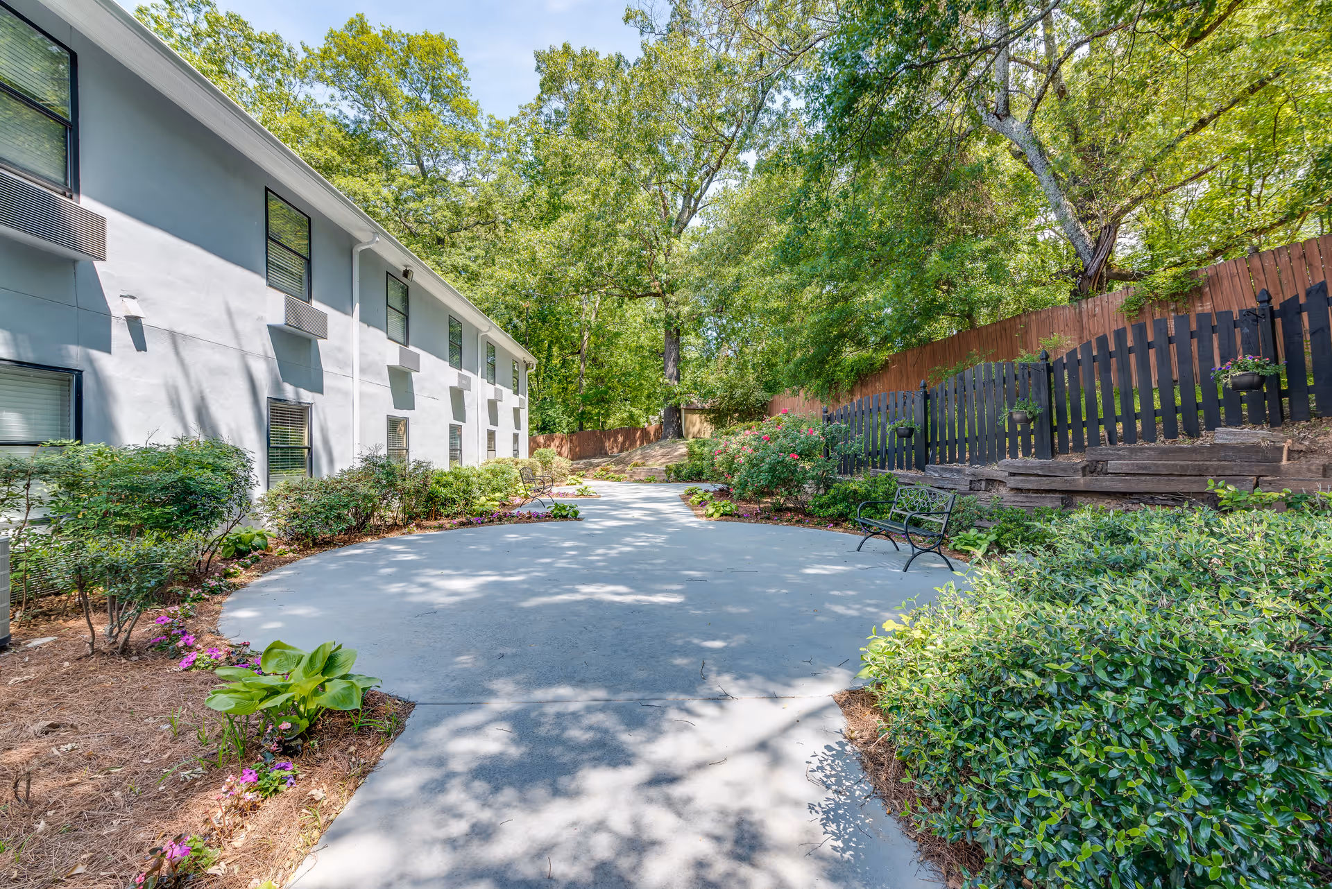 Outdoor paved pathway surrounded by green bushes, flowering plants, and trees next to a two-story building with multiple windows. There is a black metal bench on the right side near a black wooden fence with flower pots.