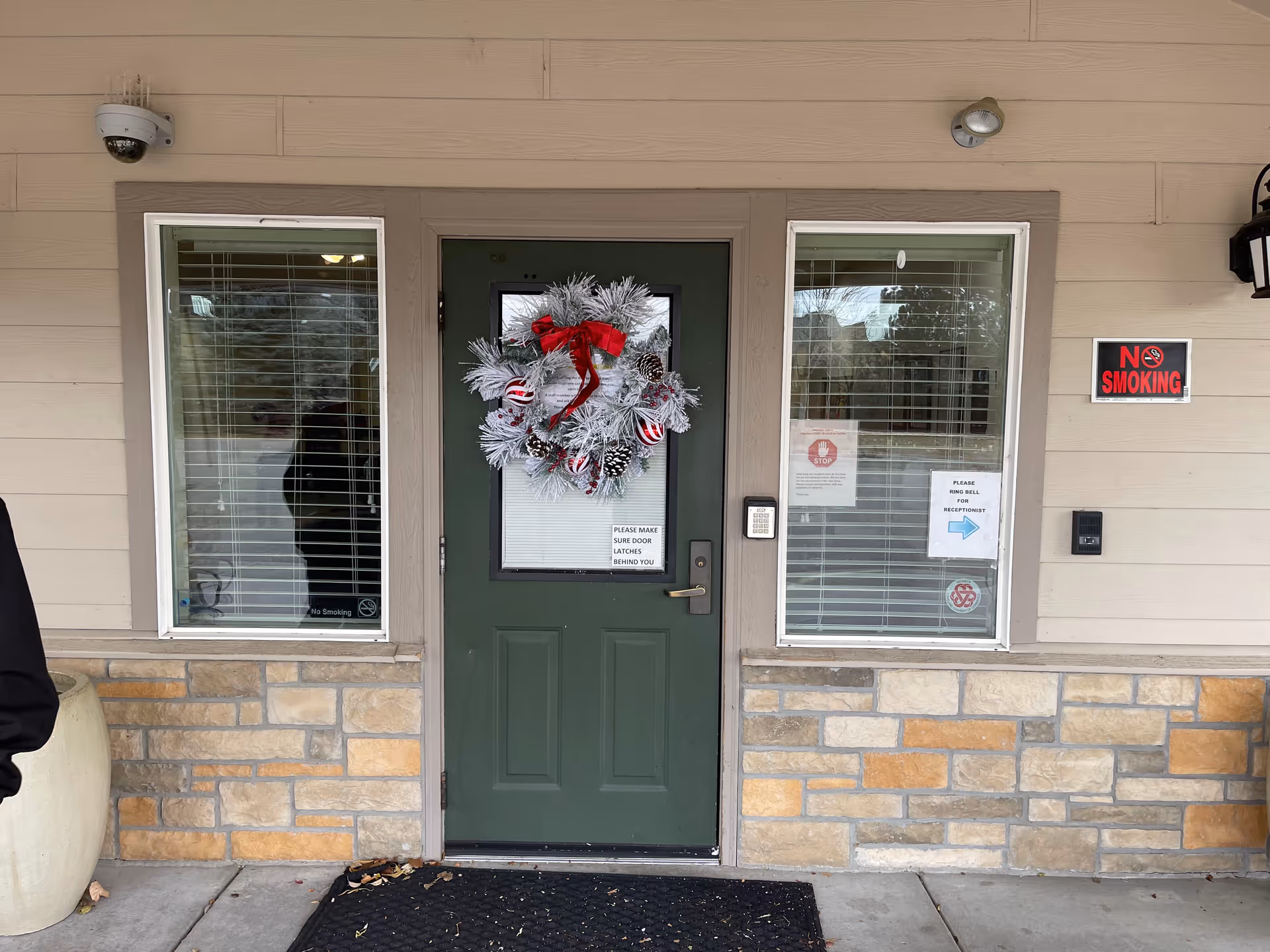 Exterior building entrance with a green door decorated with a white holiday wreath, flanked by two windows, a keypad, and a 'No Smoking' sign.