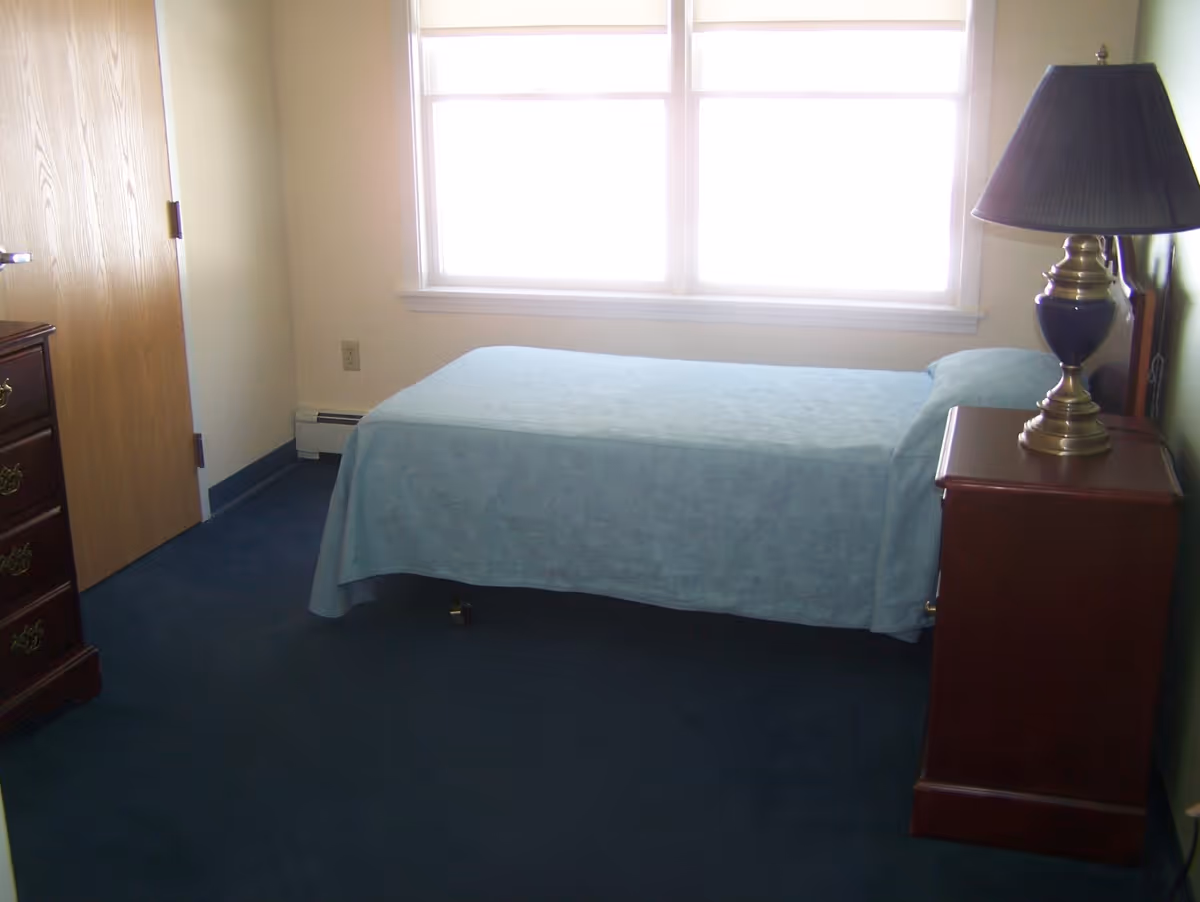 Small single bedroom with a twin bed covered in a light blue bedspread, a wooden nightstand with a lamp, dresser, and a bright window.