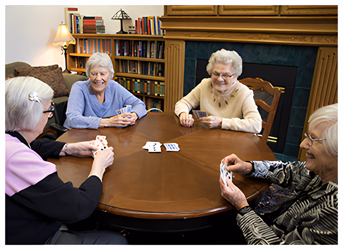 Four elderly women sitting around a wooden table playing cards in a cozy room with a fireplace and a bookshelf filled with books in the background.