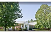 Single-story brick building with a white columned entrance, surrounded by trees and a lawn under a clear sky.
