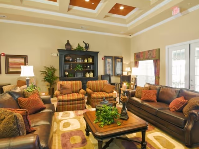 Bright communal living room with leather sofas, patterned armchairs, a central coffee table with plants, a bookcase, and a coffered ceiling.