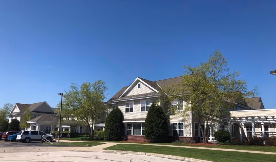 Front exterior of a two-story senior living building with parked cars, trees, and a clear blue sky.