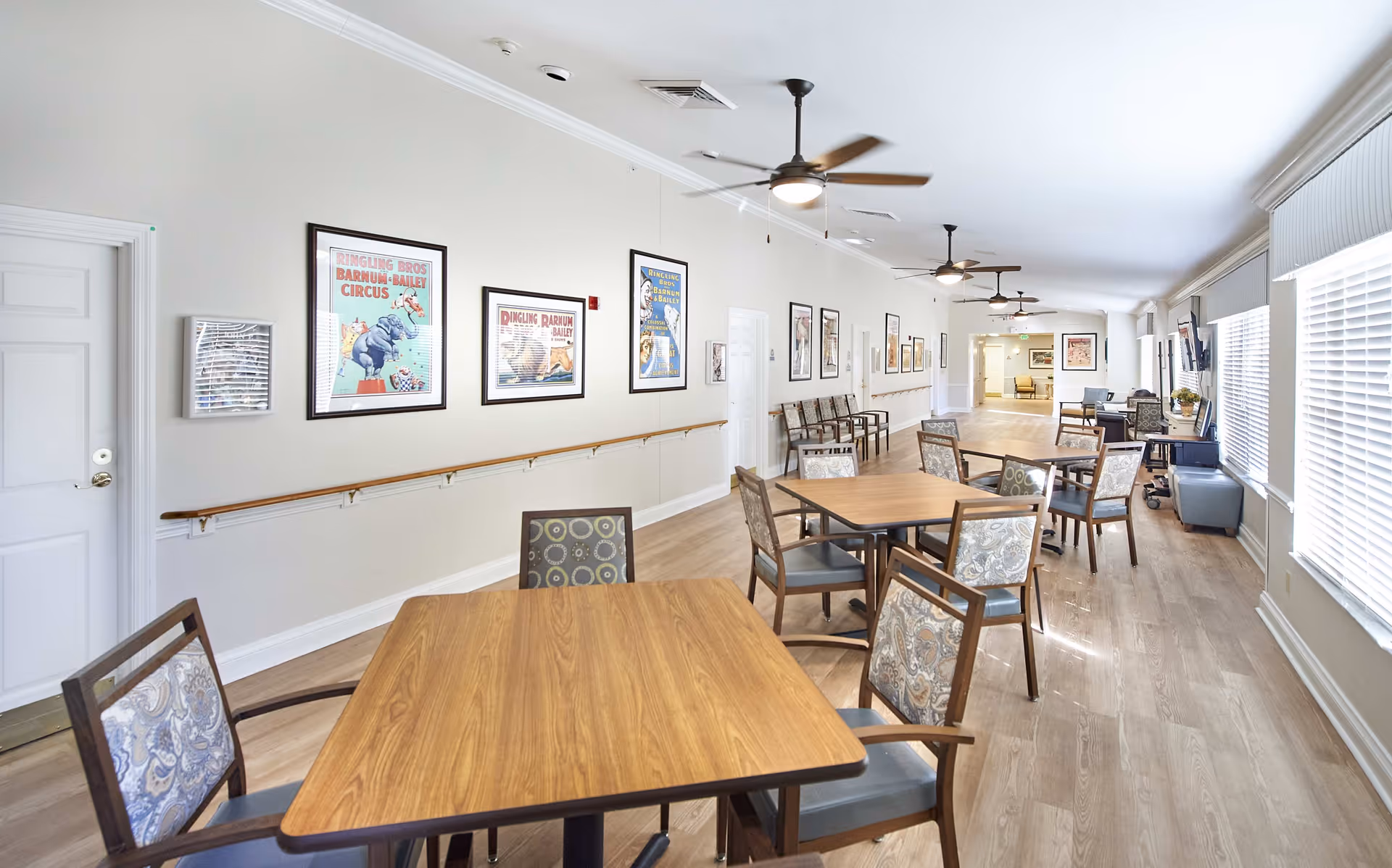 A bright and spacious common area in a senior living facility with multiple wooden tables and cushioned chairs arranged for seating. The room has light-colored walls adorned with framed vintage circus posters, ceiling fans, large windows with blinds allowing natural light, and a wooden floor. There are additional chairs lined up against the wall and a television mounted near the windows.