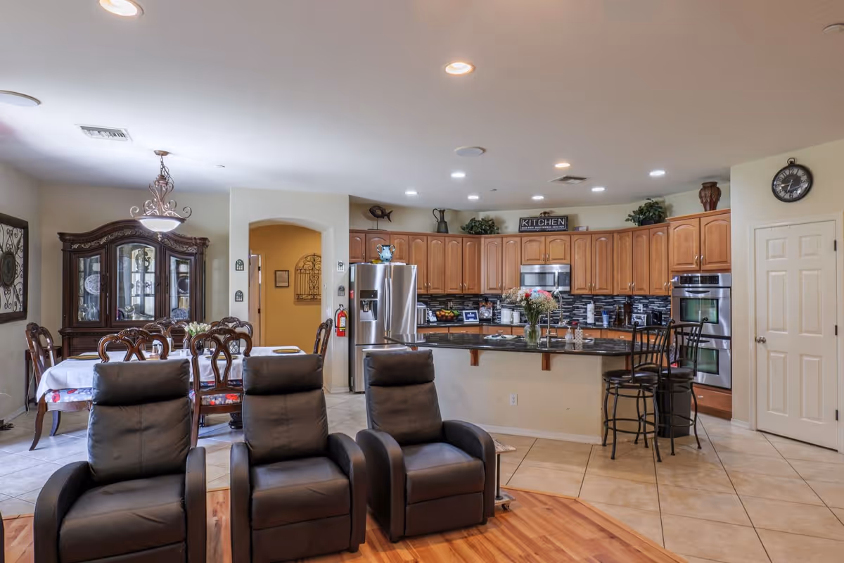 Open-plan interior showing a communal kitchen with a large island, dining table and china cabinet, and three reclining chairs in the foreground.