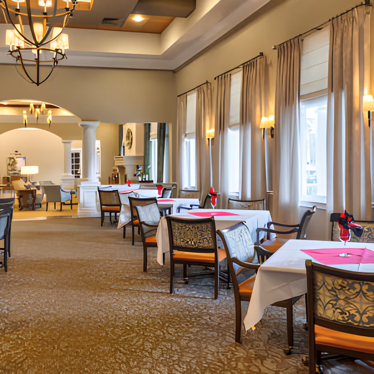 A spacious dining room with multiple tables covered in white tablecloths and red napkins folded in glasses. The room features large windows with beige curtains, wall sconces, and a chandelier hanging from a tray ceiling. The carpeted floor and elegant furniture create a warm and inviting atmosphere.