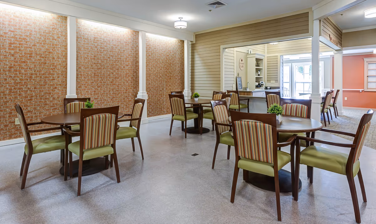 A dining area with several round wooden tables and chairs with green seats and striped backs. The room features a brick accent wall with white pillars and a small counter area labeled 'Central Cafe' in the background. The space is well-lit with ceiling lights and natural light from windows.