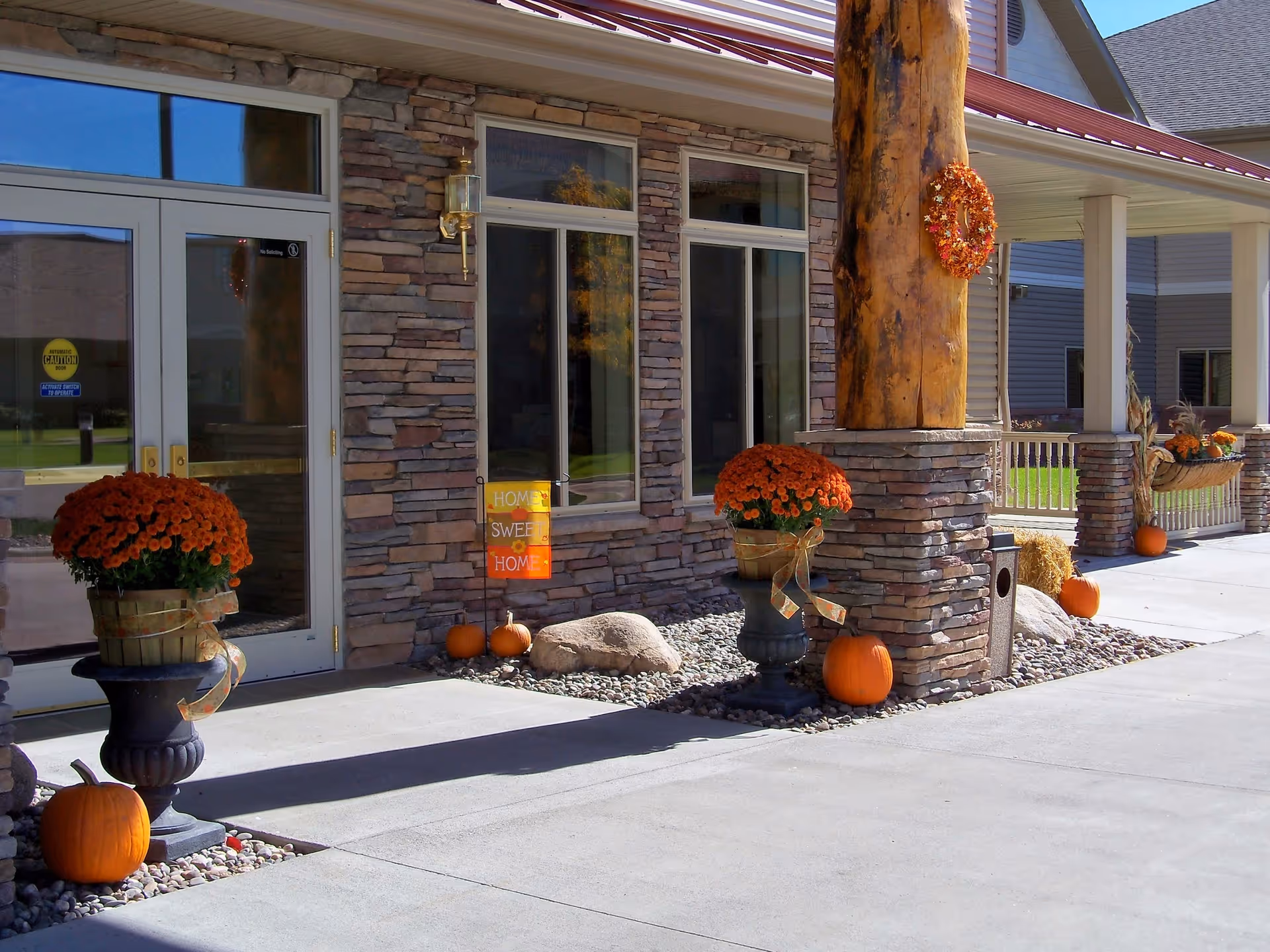 Exterior entrance of a building with stone walls and large windows. The entrance is decorated with orange flowers in pots, pumpkins, and a fall-themed wreath on a wooden pillar. A small sign near the door reads 'Home Sweet Home.'