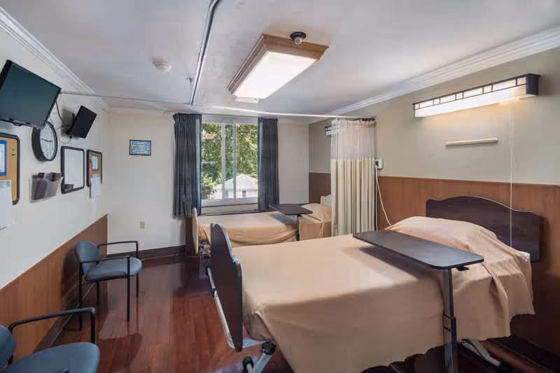 A shared patient room in a healthcare facility with two hospital beds covered in beige sheets, each equipped with an overbed table. The room has wooden flooring, two chairs, a wall-mounted television, a clock, and a window with dark curtains letting in natural light. A privacy curtain is partially drawn between the beds.