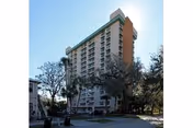 Tall multi-story residential building with balconies, surrounded by trees and a clear sky in the background.