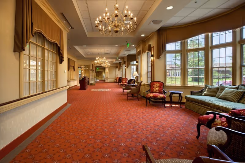 A spacious, well-lit interior hallway with large windows on the right side letting in natural light. The floor is covered with a red patterned carpet, and the ceiling features elegant chandeliers. Along the right side, there are several upholstered chairs and a green striped sofa arranged with small tables. The left side has windows with brown curtains and beige walls with decorative molding.
