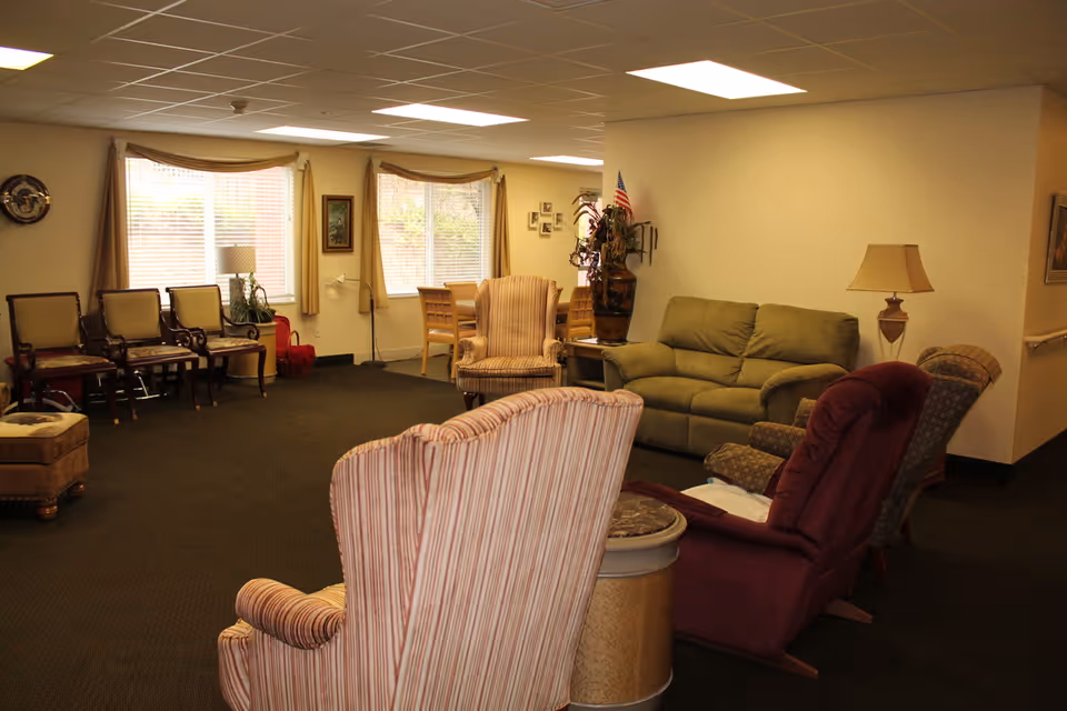 A cozy living room area in a senior living facility with various upholstered chairs, a green sofa, a lamp, and a small table. There are windows with beige curtains letting in natural light, a dining table with chairs in the background, and decorative items including a large vase with plants and an American flag.
