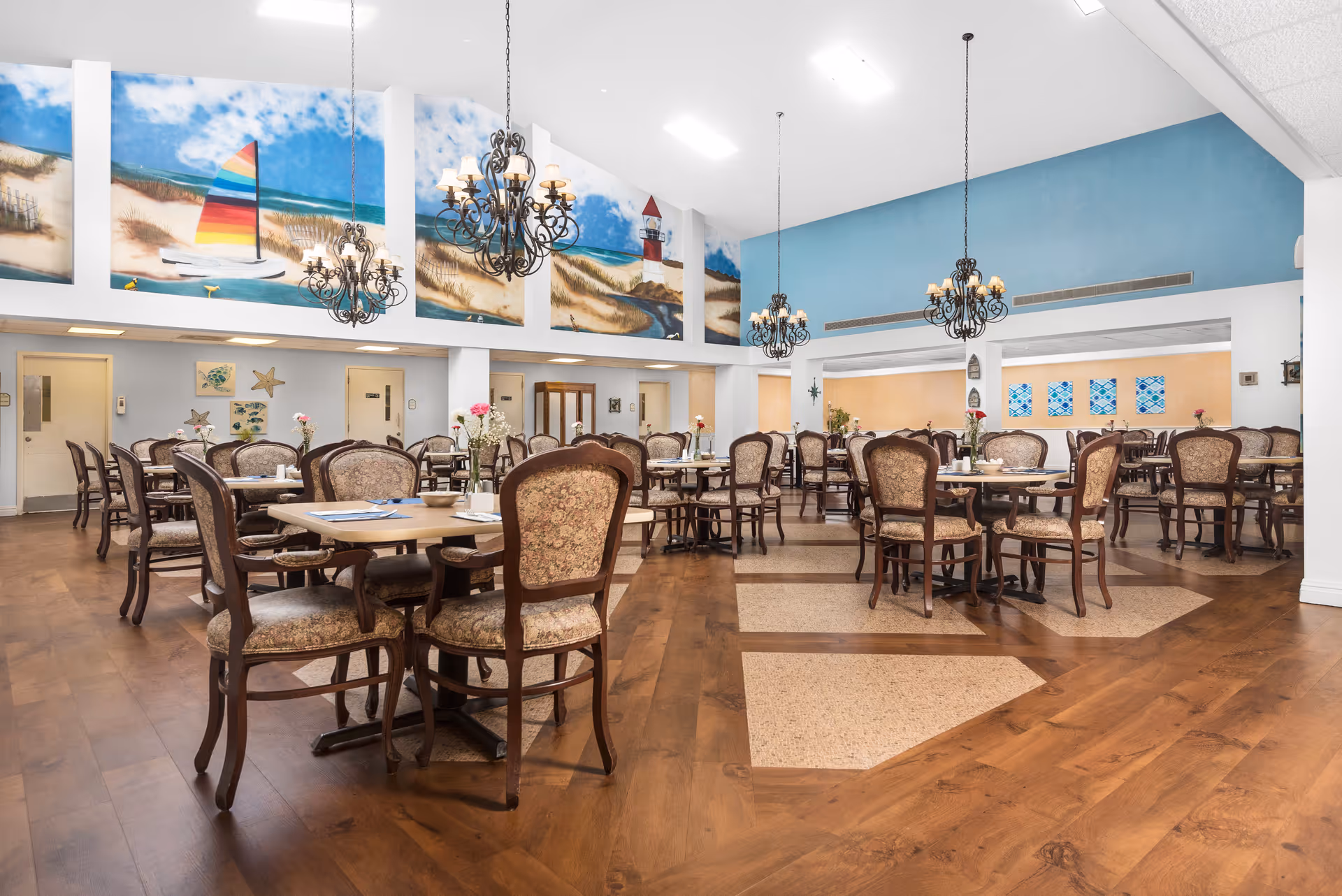 A spacious dining room with multiple round tables and ornate wooden chairs with patterned upholstery. The room features high ceilings with hanging chandeliers and large murals on the upper walls depicting a beach scene with a sailboat, sand dunes, and a lighthouse. The floor is a combination of wood and patterned tiles, and the walls are painted in soft blue and white tones.
