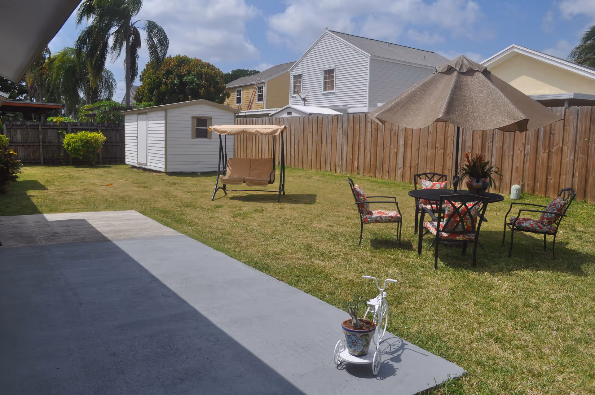 Sunny backyard with a concrete patio, lawn, storage shed, a swing, and a round patio table with umbrella and chairs beside a wooden fence.
