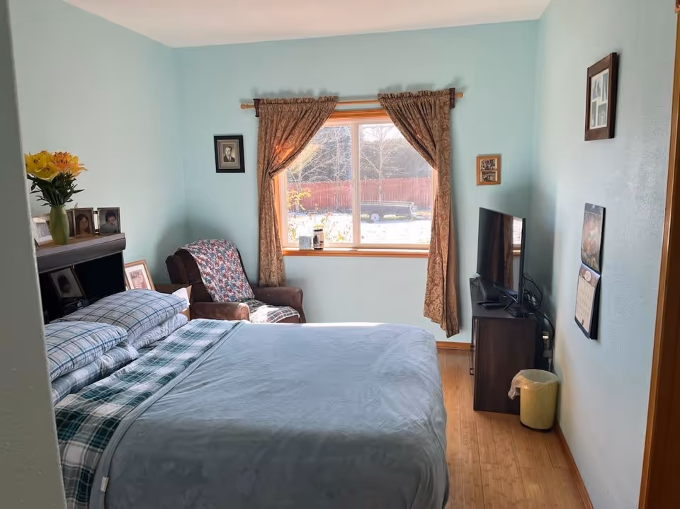 A cozy bedroom with light blue walls, a bed with plaid bedding, a brown armchair with a floral cover, a window with patterned curtains letting in natural light, a TV on a small stand, and framed photos on the walls and furniture.
