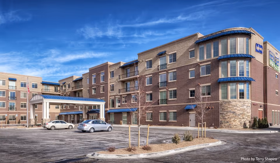 Exterior view of a multi-story assisted living and memory care facility building with a covered entrance, several parked cars, small trees, and a clear blue sky.