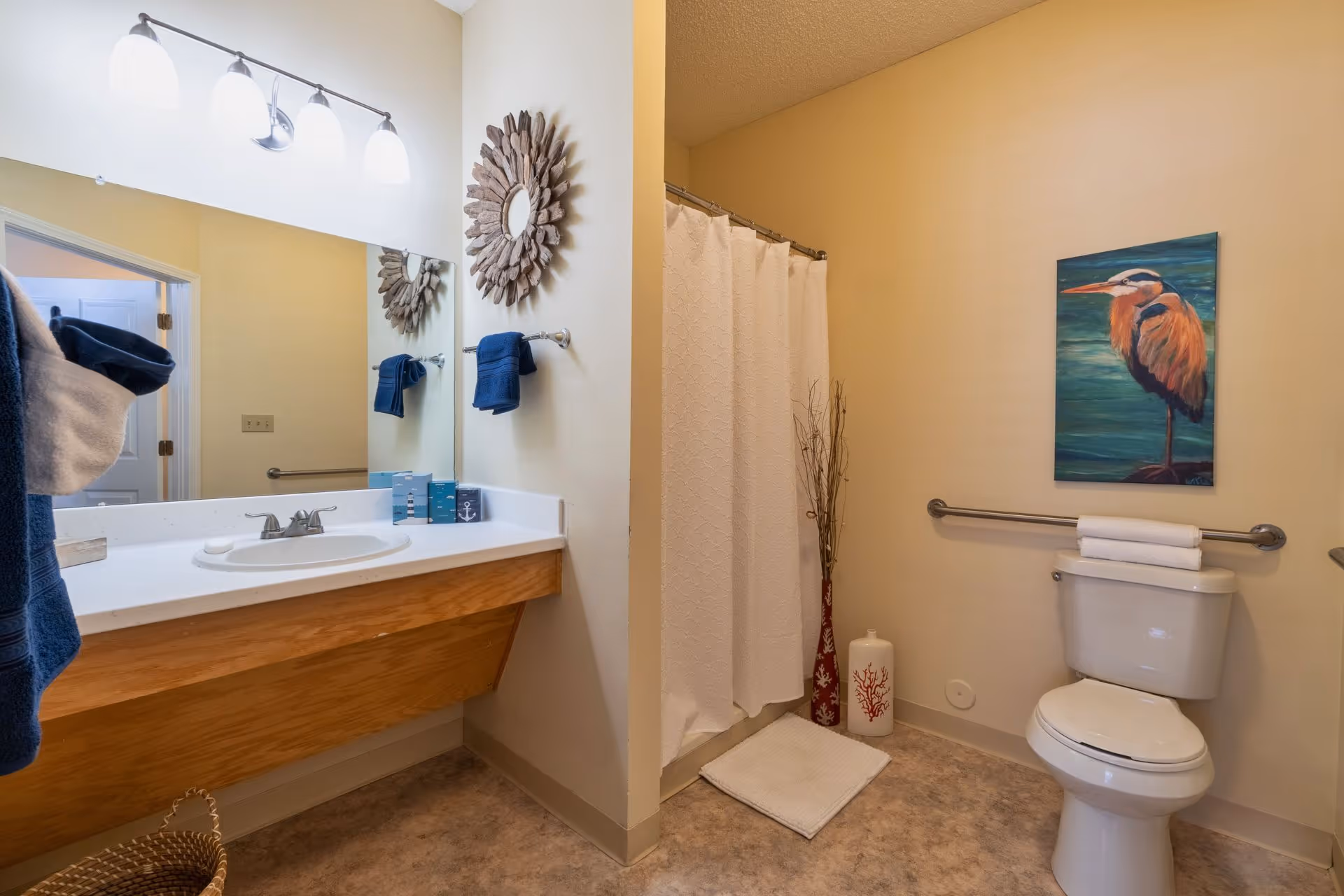 A clean and well-lit bathroom with a white sink and wooden vanity, a large mirror above the sink, a towel rack with blue towels, a shower with a white curtain, a toilet with a grab bar and folded white towels on top, a decorative painting of a bird on the wall, and decorative vases on the floor near the shower.