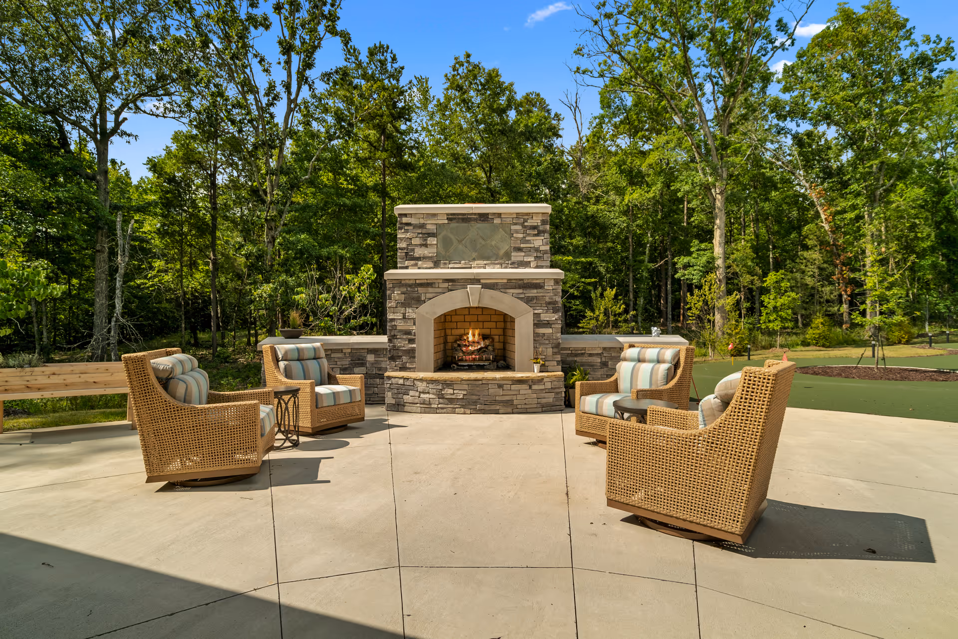 Outdoor patio area with a stone fireplace surrounded by four wicker chairs with striped cushions, set on a concrete surface with trees and greenery in the background.