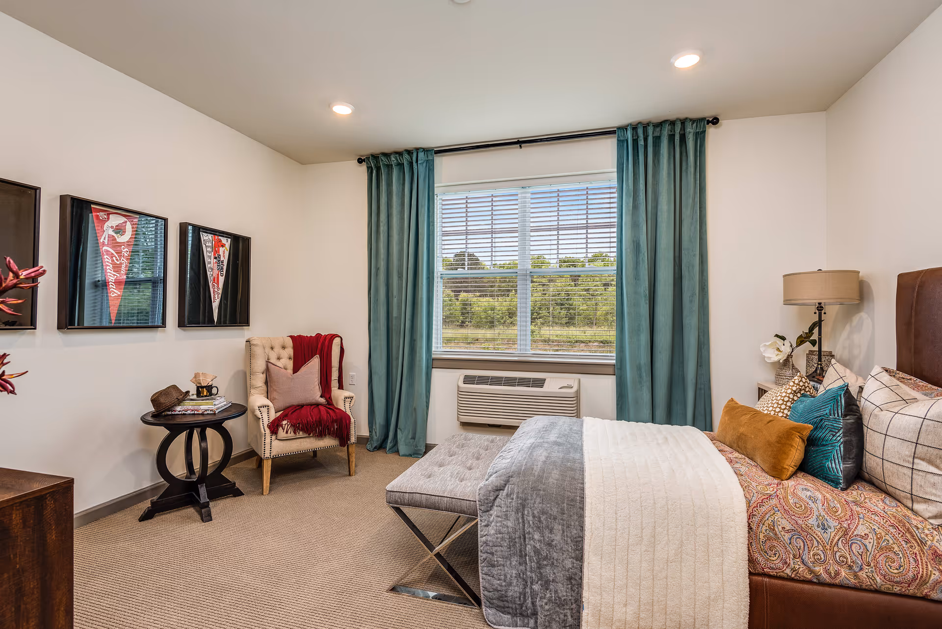 A cozy bedroom with a large window covered by teal curtains. The room features a bed with multiple pillows and a patterned bedspread, a cushioned bench at the foot of the bed, a beige armchair with a red throw blanket and pillow, a small round side table with a hat and books, and framed sports pennants on the wall.
