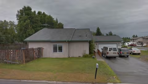 Single-story building with gray roof, driveway, fence, and parked cars.