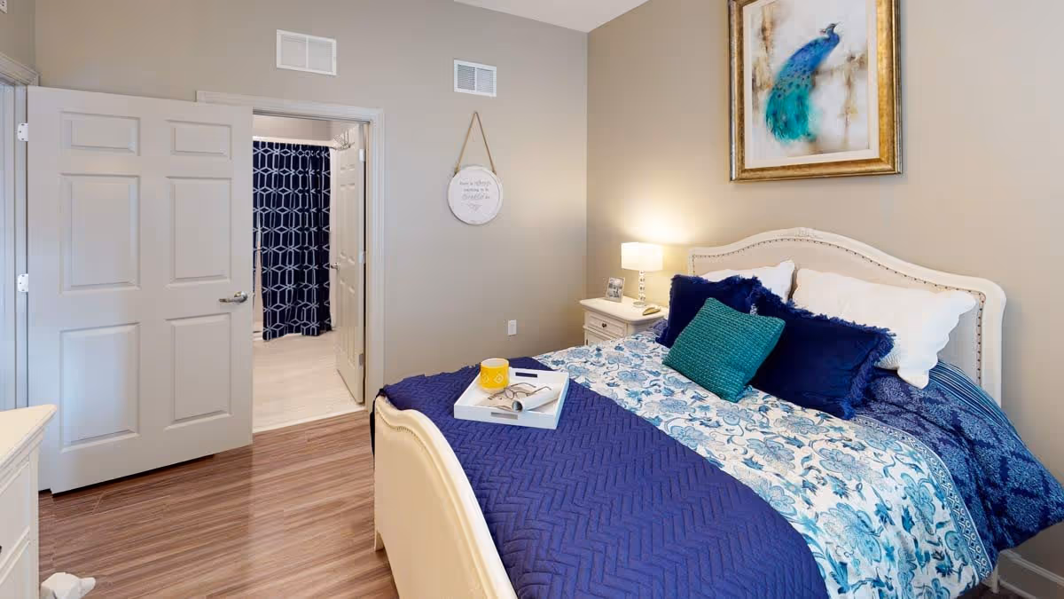 A cozy bedroom with a bed featuring blue and white floral bedding and multiple pillows. A nightstand with a lamp and a framed photo sits beside the bed. A framed peacock painting hangs on the wall above the bed. The bedroom has wooden flooring and an open door leading to a bathroom with a blue patterned shower curtain.