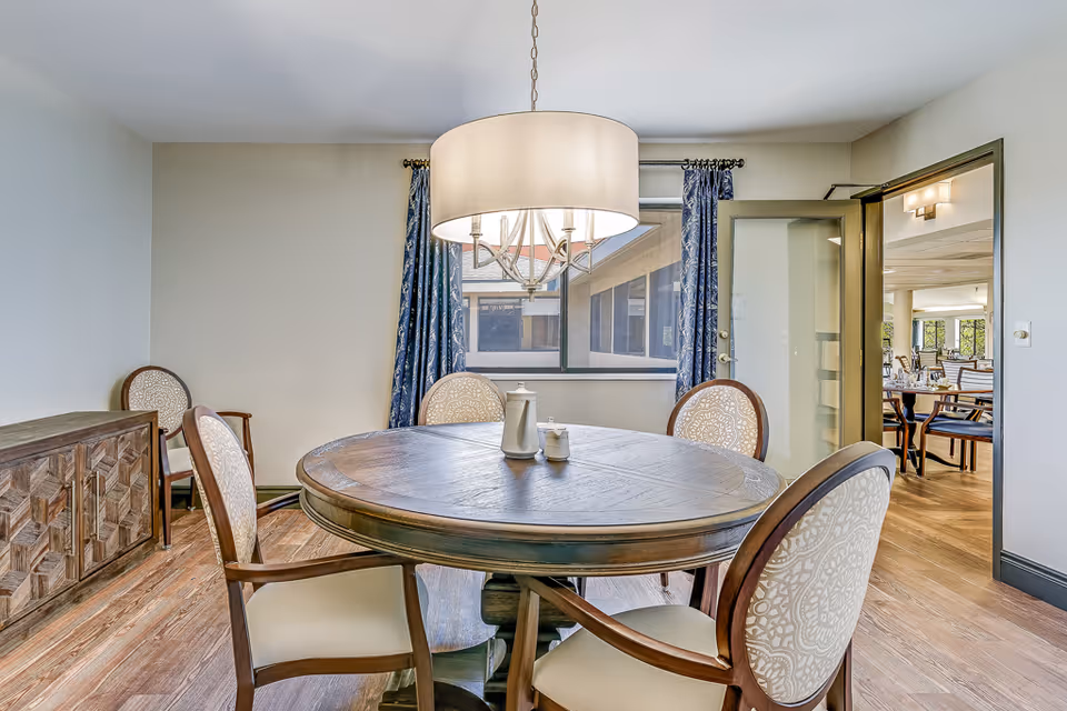 Round wooden dining table with four upholstered chairs under a drum pendant light in a bright dining room with wood floors and a sideboard.