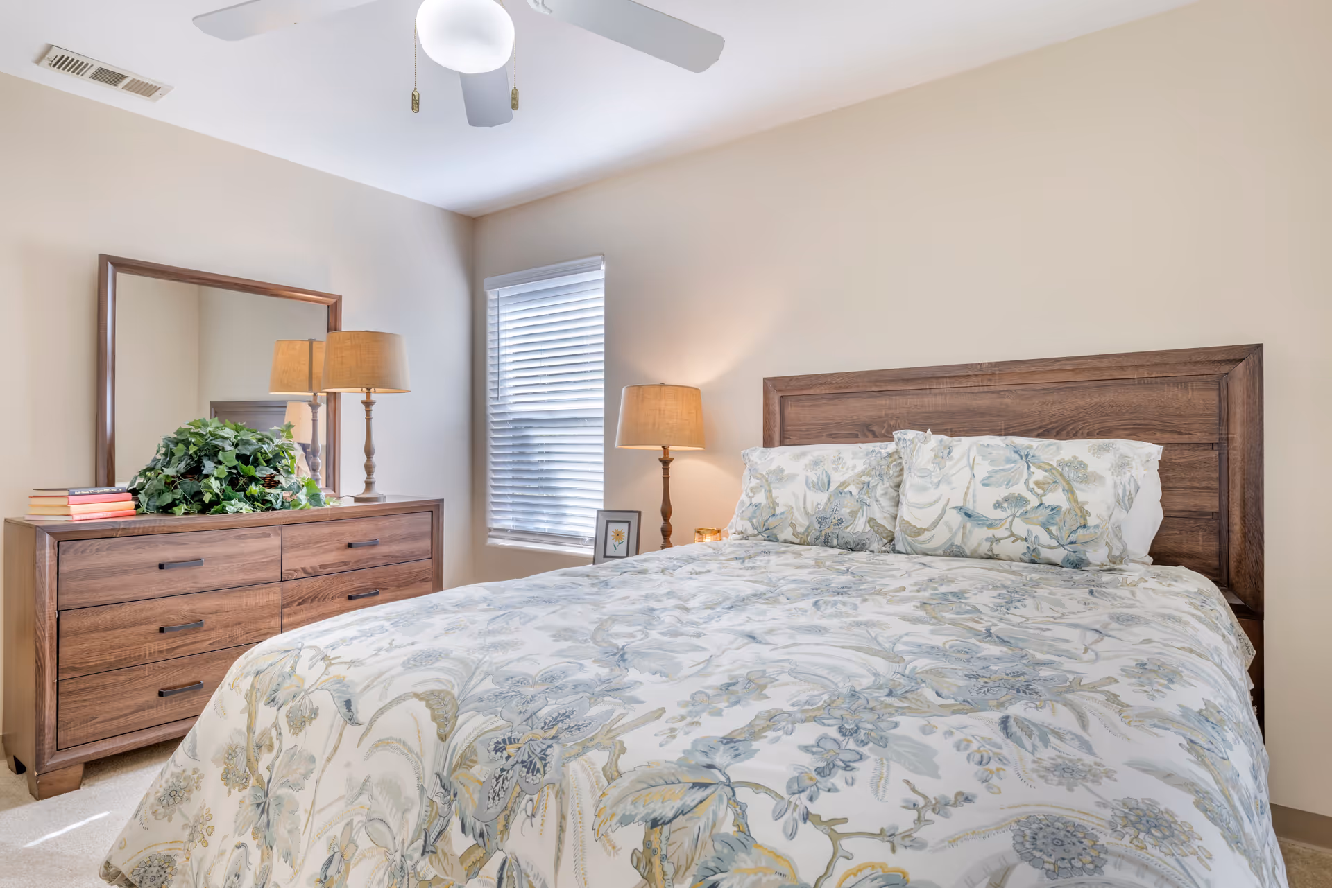A bright bedroom with a wooden bed frame and floral patterned bedding. There is a wooden dresser with a large mirror, a table lamp, a stack of books, and a green plant on top. A window with white blinds is next to the bed, and a ceiling fan is visible above.