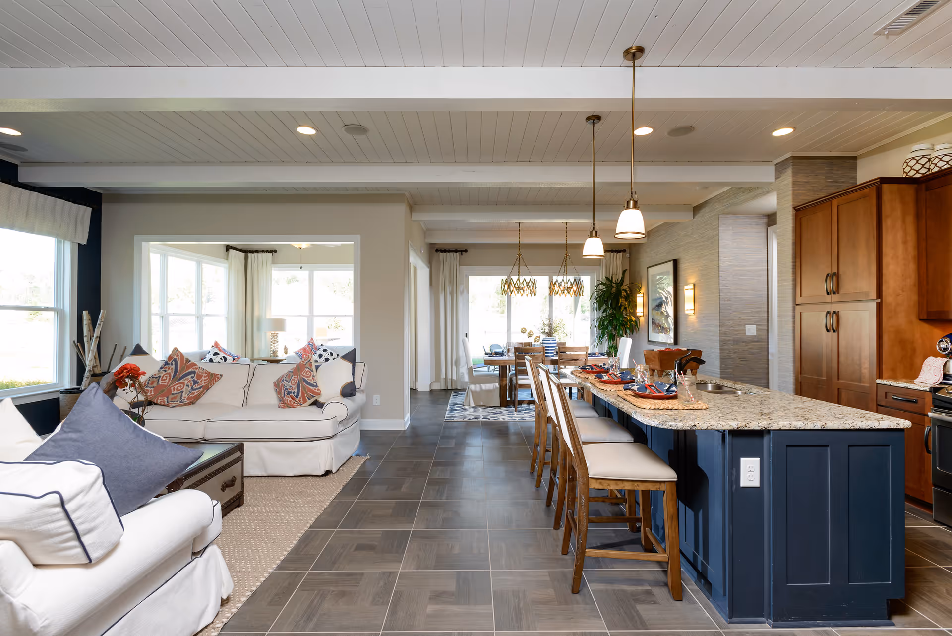 Open concept interior of a senior living facility showing a living room with white sofas and colorful pillows on the left, a kitchen area with a granite countertop island and wooden bar stools on the right, and a dining area with a wooden table and chairs in the background. The space is well-lit with large windows and pendant lights hanging from the ceiling.
